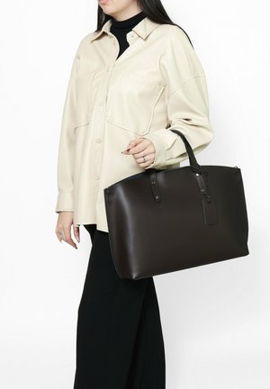 Borsa a tracolla in pelle marrone con forma strutturata, manici superiori e un piccolo dettaglio con etichetta; abbinata a una camicia oversize beige chiaro.