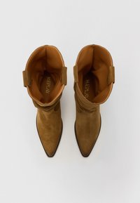 Brune ankelstøvler i ruskind med spidse tæer, slaskede design og dekorative løkker øverst. Foret med lysebrunt stof.