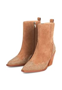 Botas de tobillo de ante en tono marrón claro con punteras afiladas, presentando bordes ondulados y detalles de tachuelas en la zona de los dedos. Tacón de madera grueso. Cierre con cremallera lateral.
