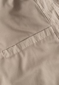 Tessuto beige strutturato con un motivo a rete, caratterizzato da una tasca piatta cucita e un pannello liscio a contrasto in una tonalità più chiara.