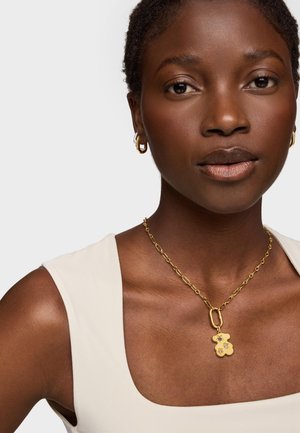 Collar de cadena dorada con un colgante en forma de oso que presenta acentos florales. El modelo lleva una camiseta sin mangas de color crema claro.