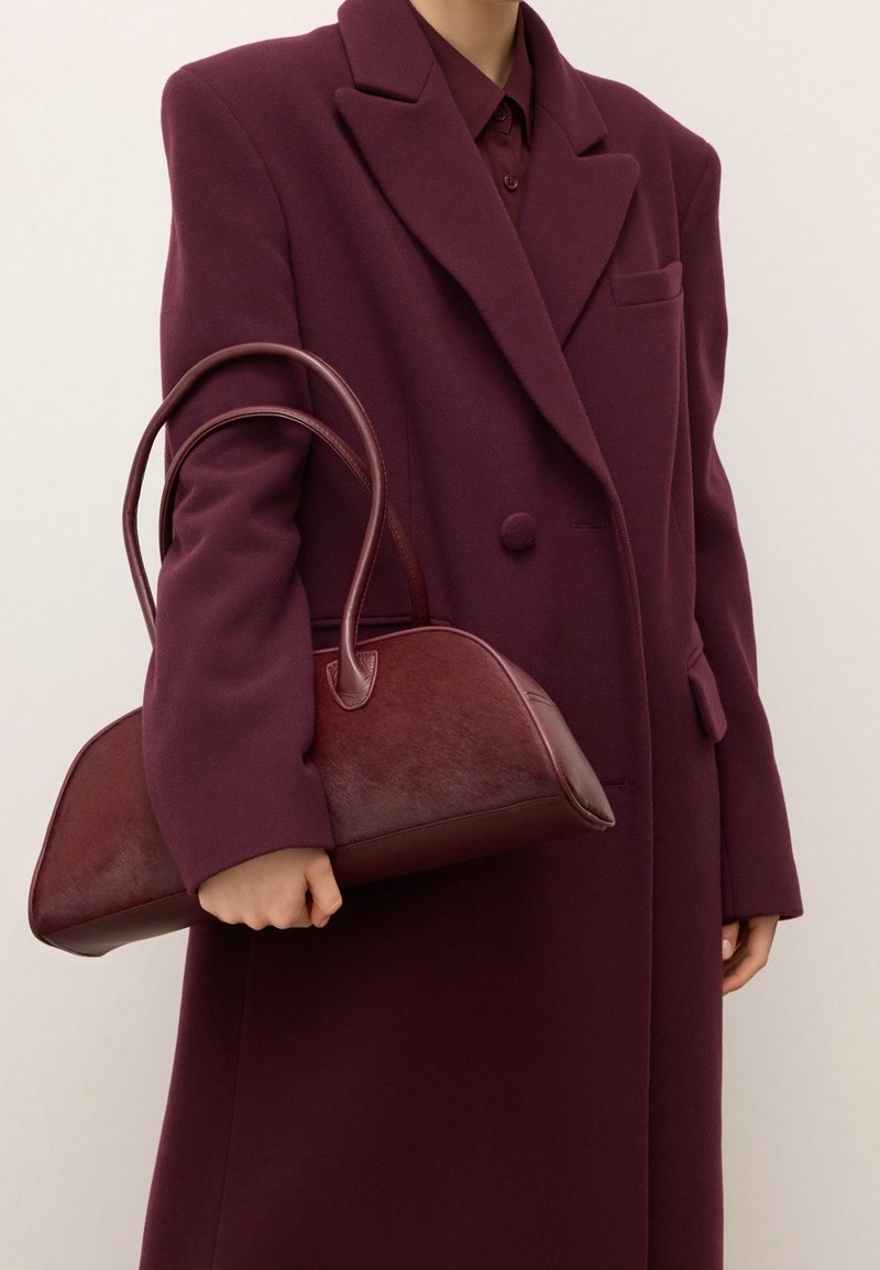 Personne portant un manteau et une chemise bordeaux tenant un sac à main en cuir rouge foncé avec des doubles poignées au niveau de la taille.