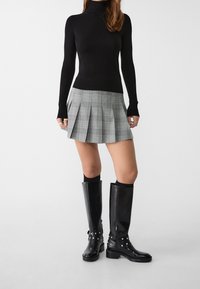 Czarny sweter z golfem połączony z szarym, w kratę, plisowanym mini spódnicą. Modelka nosi czarne, skórzane kozaki do kolan z dekoracyjnym wykończeniem.