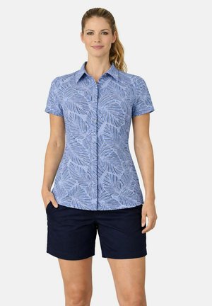 Donna che indossa una camicia blu a maniche corte con fantasia e bottoni e pantaloncini blu scuro, in piedi con una mano in tasca davanti a uno sfondo semplice.