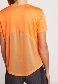 Oranžové sportovní tričko s prodyšnou síťovanou částí na zádech. Má krátké rukávy, zaoblený lem a malou bílou značku na zádech.