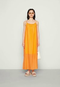 Mulher vestindo um vestido maxi laranja sem mangas, sandálias de plataforma bege, óculos de sol com lente laranja, brincos de argola, segurando uma pequena bolsa branca.