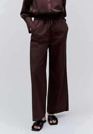 Osoba nosząca luźne ciemnobrązowe spodnie ze ściągaczem w talii i czarne sandały z odkrytymi palcami, stojąca na jednolitym tle.