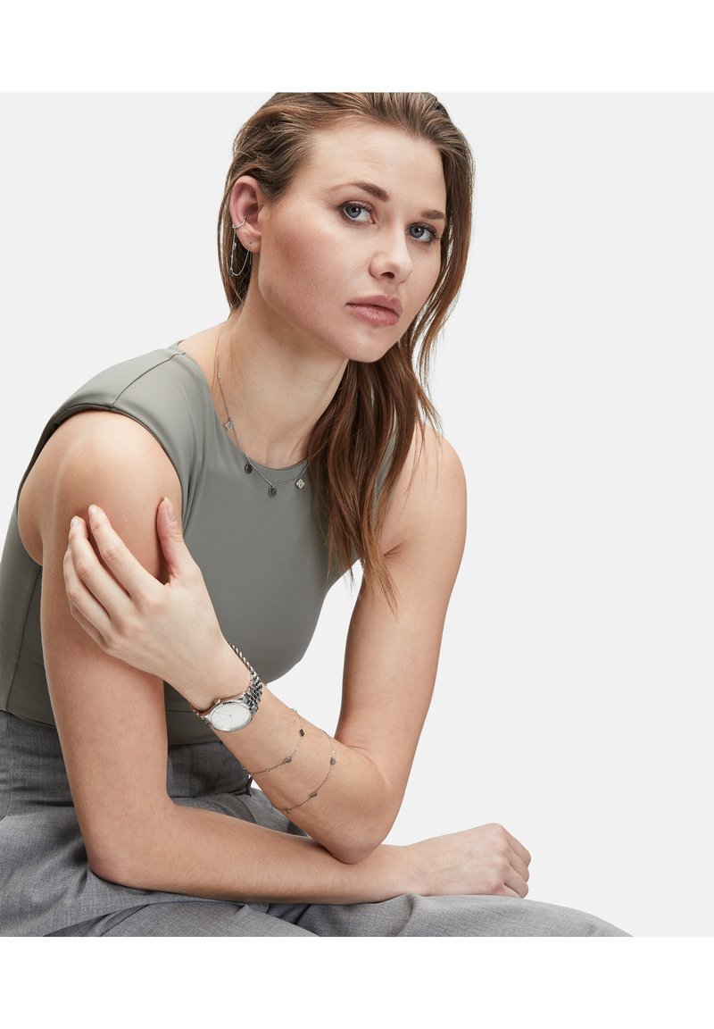 Sølvur med rund skive, grå top, lagdelte sølvhalssmykker og et gråt bukser, der fremhæver elegante materialer og moderne design.