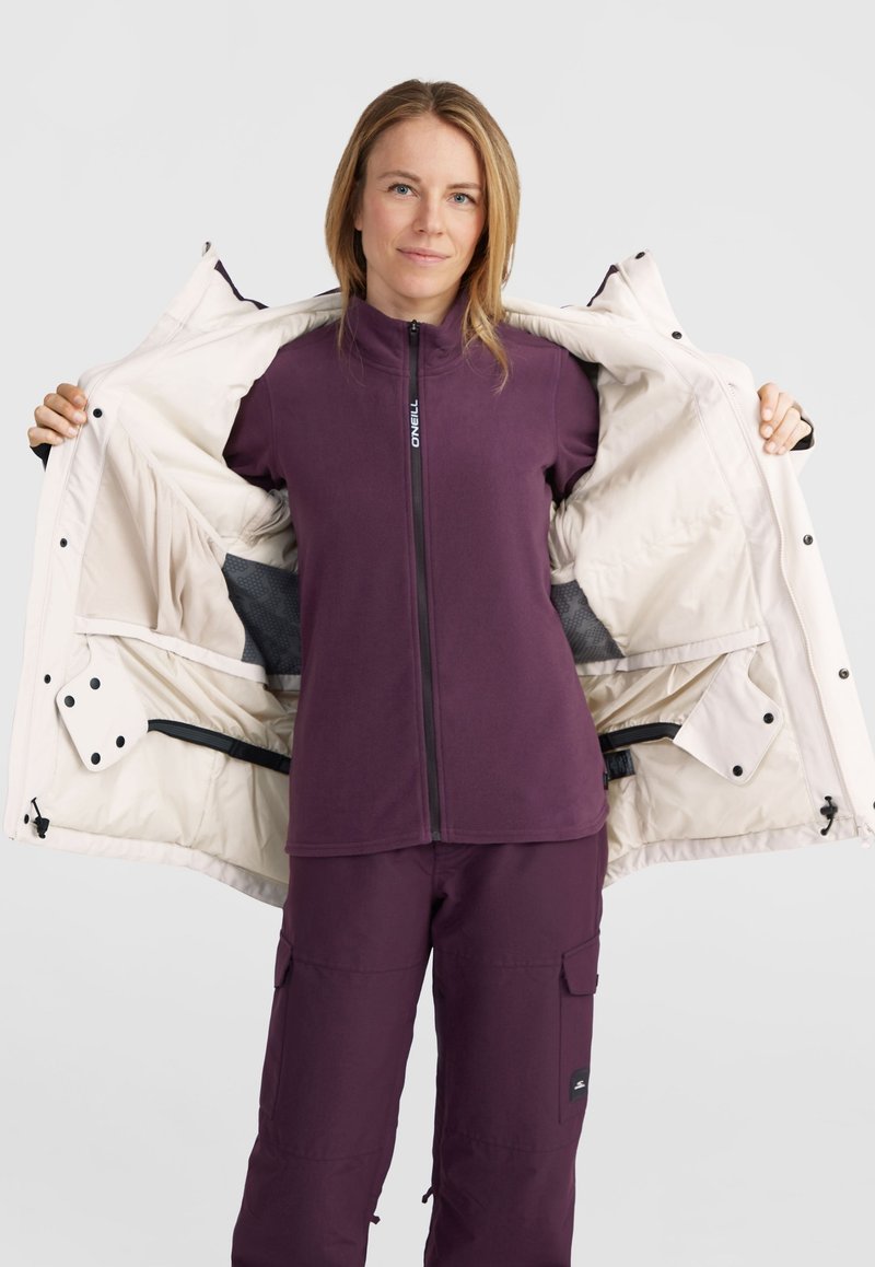 Isolierte weiße Jacke mit Druckknöpfen, offen über einem lila Fleece-Oberteil getragen. Das Modell trägt außerdem passende lila Hosen mit Cargo-Taschen.