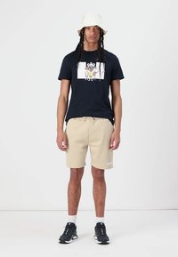 Camiseta negra con un diseño gráfico, pantalones cortos beige y zapatillas deportivas oscuras. El conjunto incluye un sombrero tipo cubo y está frente a un fondo liso.