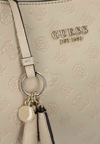 Borsa beige con motivo logo in rilievo, caratterizzata da un anello in metallo dorato e un'etichetta, superficie testurizzata e profili neri a contrasto.