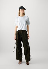 Weißes T-Shirt mit puffärmel, kombiniert mit lockeren, dunkelgrünen Cargo-Hosen mit Seitentaschen und verstellbaren Bändern. Schwarze Mütze und Sandalen.
