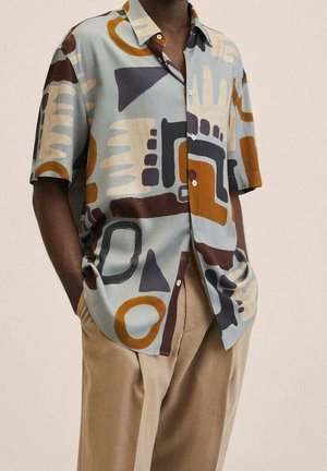 Chemise à manches courtes à boutons avec des motifs géométriques abstraits en bleu, marron, beige et noir, associée à un pantalon beige.