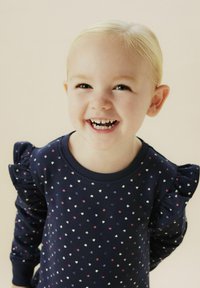 Suéter marino con un pequeño patrón de corazones multicolores, con mangas rizadas, cuello redondo y textura suave. Niño sonriente, con cabello corto y rubio.