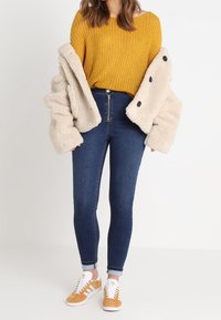 Knitted mustard sweater, cream faux fur coat with black buttons, dark blue skinny jeans, and tan sneakers with white accents.