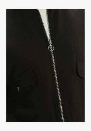 Chaqueta negra con cremallera, hecha de un material suave, que presenta una cremallera plateada y bolsillos en el pecho con botones. Textura suave con un diseño minimalista.