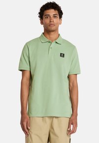 Ljusgrön polo tröja i bomull med klassisk krage, korta ärmar och en svart logotyp på vänster bröst. Slät textur.