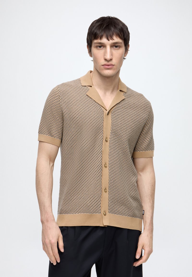 Jeune homme portant une chemise beige à motifs à manches courtes et un pantalon noir sur un fond clair uni.