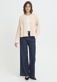 Cardigan roz deschis cu decolteu rotund și șase nasturi, purtat peste un top alb și asortat cu blugi evazați de culoare albastru închis.