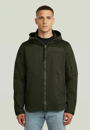 Mann trägt eine dunkelgrüne Kapuzenjacke mit Frontreißverschluss und Taschen, kombiniert mit hellblauen Jeans, vor einfarbigem Hintergrund stehend.