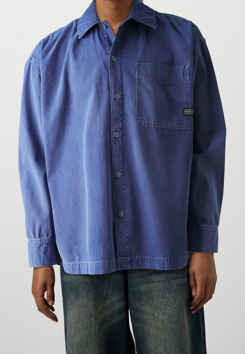 Långärmad, skjorta med knappar i dämpad blå färg. Tillverkad av mjukt material med en enda bröstficka och en avslappnad passform. Har klassisk krage och manschetter.