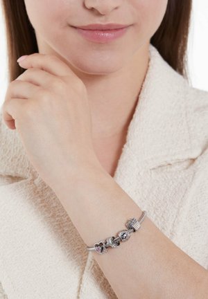 Silberne Charm-Armbanduhr mit Herz-, Cupcake- und Perlenanhängern; zeichnet sich durch filigrane Details und Oberflächen aus; am Handgelenk vor einem strukturierten Stoff-Hintergrund getragen.