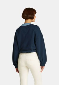 Felpa oversize blu navy con spalle scese, polsini a costine e un dettaglio a forma di V sul retro, abbinata a jeans bianchi slim-fit.