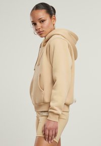 Felpa beige con cappuccio realizzata in morbido tessuto, dotata di tasca frontale e polsini a coste. Abbinata a pantaloni corti aderenti coordinati.