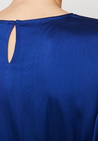 Haut bleu roi avec une texture côtelée, présentant un détail dos ajouré et un col rond, fabriqué en tissu lisse et léger.