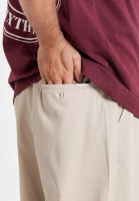 Beige trousers with a side pocket and a visible label. A maroon t-shirt covers part of the pocket. Hand is inserted into the pocket.