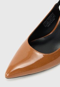 Καφέ παπούτσι γόβας από πατέντα, με μυτερή μύτη και γυαλιστερή επιφάνεια, διαθέτοντας χαμηλή μπροστινή κοπή και μαύρη εσωτερική επένδυση.