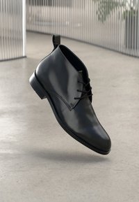 Μαύρη δερμάτινη μπότες στον αστράγαλο με λεία επιφάνεια, στρογγυλή μύτη και κορδόνια. Διαθέτει μια μικρή θηλίτσα και χαμηλό τακούνι. Το φόντο είναι μινιμαλιστικό.