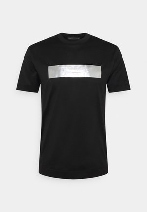 Musta lyhythihainen t-paita, jossa on metallinhohtoinen hopeinen suorakaiteen muotoinen logopatch, jossa on tyylitellyt kirjaimet "E" ja "A" sekä kotkaembleemi keskellä.