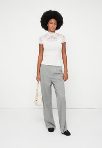 Top blanco de encaje con cuello alto y mangas cortas, combinado con pantalones de rayas grises y zapatos planos negros. Lleva un pequeño bolso beige.