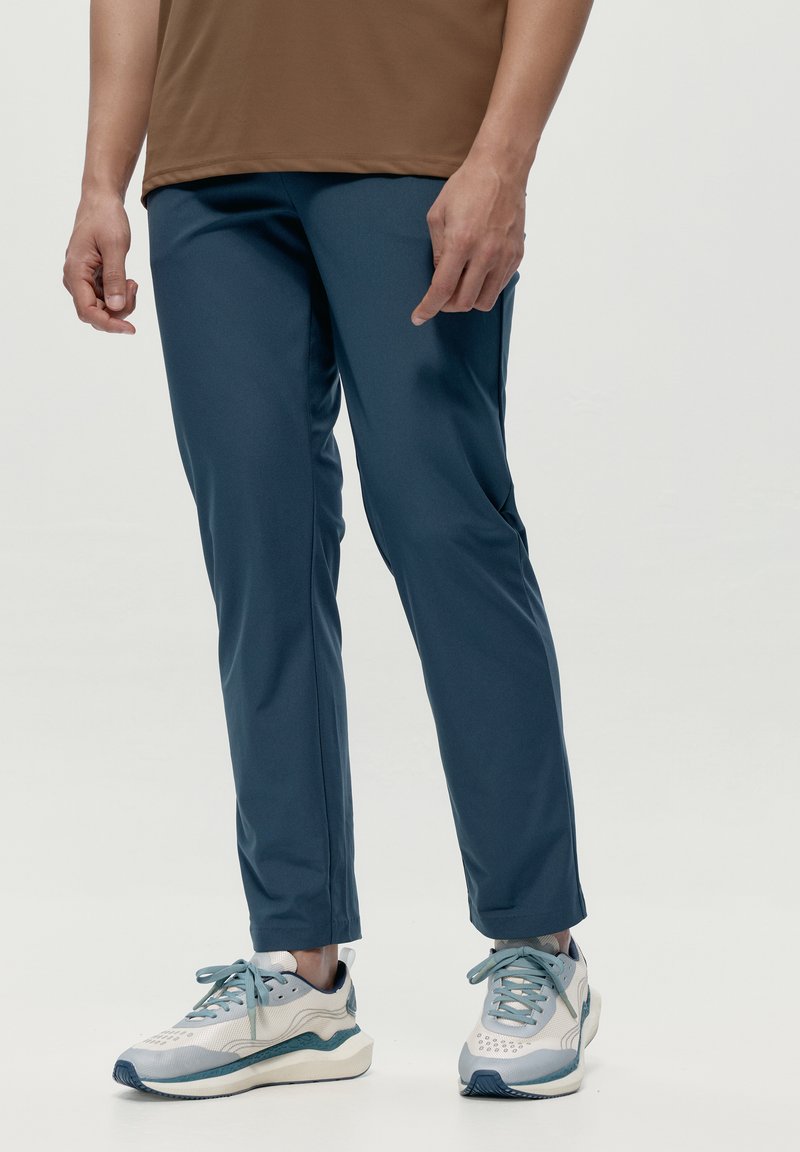 Modré športové nohavice s hladkou textúrou, zúženým strihom a bočnými švami. Nosené s svetlosivými teniskami s modrými akcentmi.