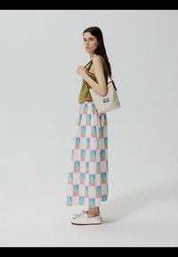 Femme portant un débardeur olive, une longue jupe à motifs, des chaussures blanches, tenant un sac à bandoulière beige, debout devant un fond clair uni.