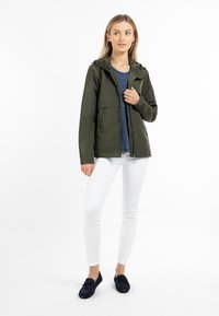 Giacca leggera verde scuro con cappuccio, chiusura con zip e due tasche laterali, indossata sopra una camicia a righe blu navy e bianche e pantaloni bianchi.
