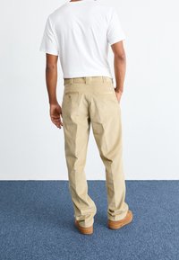 Béžové chino kalhoty se střihovým designem rovného nohavice, dvěma zadními kapsami a textilní štítkem poblíž pasu. Spárováno s jednoduchým bílým tričkem a hnědými botami.
