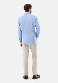 Camicia azzurro chiaro a maniche lunghe in cotone, con orlo curvo, abbinata a pantaloni beige e scarpe nere, vista da dietro.