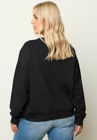 Felpa nera con vestibilità rilassata, polsini e orlo a costine. Tessuto morbido, design minimalista, indossata con jeans blu chiaro.