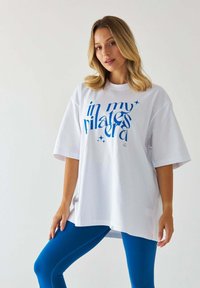 Biała, oversize'owa koszulka z niebieskim napisem "w mojej erze pilatesu", noszona z dopasowanymi niebieskimi legginsami. Bawełniany materiał, krótki rękaw i luźny krój.