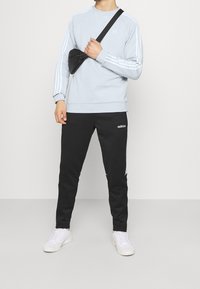 Světle modrá mikina s bílými pruhy, černé sportovní kalhoty s bílým logem a bílé tenisky. Obsahuje malou černou crossbody tašku.