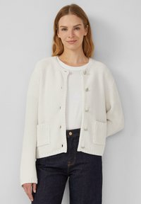 Cardigan blanc texturé avec un col rond, des poches avant et une fermeture par boutons, associé à un t-shirt blanc et un jean noir.