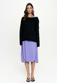 Czarny, oversize, długi rękaw top zestawiony z fioletową spódnicą balonową. Czarny sandały dopełniają stylizację. Czerwone kolczyki w kształcie kwiatów dodają szczegółu.