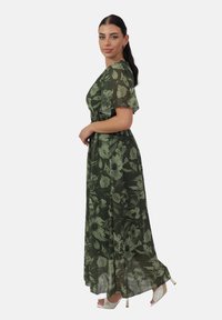 Maxikleid aus durchscheinendem grünem Stoff mit floralem Muster, kurzen Flatterärmeln, tailliertem Bund und einem fließenden Rock. Kombiniert mit Pumps.
