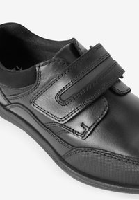 Chaussure en cuir noir à profil bas, dotée de deux sangles Velcro, côtés texturés et coutures visibles. Fini lisse.