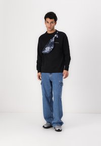 Zwarte sweatshirt met logo, blauw tie-dye schoudertas, lichtblauwe wide-leg jeans en zwarte en zilveren sneakers. Minimalistische achtergrond.