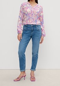 Blouse légère à manches longues de couleur rose pâle avec un imprimé botanique bleu et violet ; portée avec un jean bleu retroussé et des talons roses à brides.