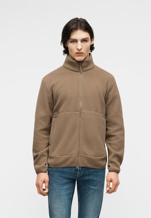 Joven con una chaqueta de vellón marrón con cremallera completa y jeans azules, de pie contra un fondo claro y liso.