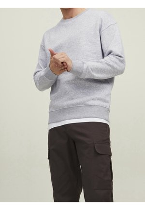 Persona con un sudadera gris claro sobre una camisa blanca y pantalones cargo oscuros, manos entrelazadas en la cintura, contra un fondo liso.